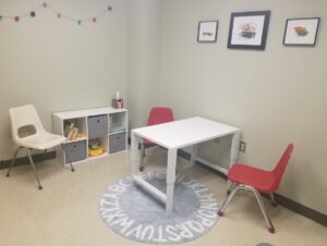 This is a picture of the LangLitLife laboratory room with child sized furniture including a white table, two red chairs, a rug with the alphabet on it and small bookcase with books and toys. On the wall are three pieces of colorful artwork of a caterpillar, bird and boat with fireworks.